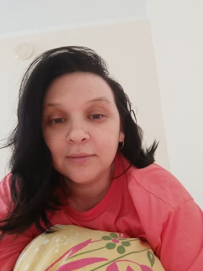 A medium shot from a slightly high angle shows a woman with dark hair and light skin. She is looking directly at the camera. She is wearing a pink t-shirt and a gold hoop earring in her left ear. Her head is resting on a pillow with a yellow floral pattern. The background is a plain white wall. This image was taken indoors, likely in a bedroom, in Krasnyi Pakhar', Russia.