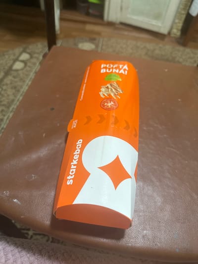 The image shows a food container from Starkebab, an establishment in Corlăteni, Moldova. The container is orange and white, with text in Romanian. It features the Starkebab logo and the phrase "POFTĂ BUNĂ!" (Bon appétit!). The container is placed on a brown, textured surface, possibly a table. The background is blurred but suggests an indoor setting with wooden flooring and furniture. There are no people visible in the image.