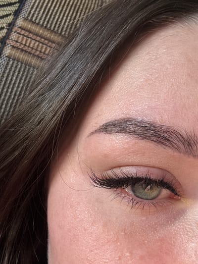 This is a close-up, angled photograph of a person's eye and eyebrow. The subject has fair skin with visible pores. Their eye is open, revealing a green iris. Long, dark eyelashes frame the eye, and a dark eyebrow arches above it. The person's dark hair is visible in the foreground, partially obscuring the lower part of the image. The background appears to be a striped fabric, possibly a blanket or upholstery. The lighting suggests an indoor setting, and the focus is sharp on the eye and eyebrow, with the hair slightly out of focus. There are no visible text elements in the image. The overall impression is one of a personal, intimate portrait focused on facial features. The crops highlight different areas of the face and hair, emphasizing the texture of the skin, the individual hairs of the eyebrow and eyelashes, and the weave of the fabric.