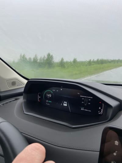 The image shows the interior of a car, specifically the dashboard and the view through the windshield, likely taken from the driver's perspective. The car is in motion, indicated by the speed of 149 km/h displayed on the digital dashboard. The weather appears to be overcast and rainy, with raindrops visible on the windshield. The dashboard displays various information including tire pressure (2.4 bar), engine RPM (2.9 x1000r/min), and the current gear (D for Drive, NORMAL mode). Outside the car, a green, grassy roadside with trees can be seen. The location is identified as Поварёнки, Russia based on the context. A hand is visible in the lower left corner, suggesting the driver is operating the vehicle. A portion of a smartphone or secondary display is visible in the lower right corner.