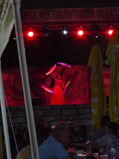 This image captures an outdoor performance at night, possibly a festival or a show. The scene is illuminated by vibrant red stage lights, creating a dramatic atmosphere. In the foreground, a performer in a bright orange dress is juggling several glowing, colorful objects, which appear to be batons or props. They are positioned in front of a dark backdrop with a textured, abstract pattern illuminated in shades of red.

To the left, a large sign with Cyrillic text is partially visible, reading "OPEN FESTIVAL LIVE WATER." The sign is mounted on a metal truss structure, also lit by red lights. In the background, yellow awnings or umbrellas are draped overhead, suggesting a sheltered area or seating for an audience. Beyond the performance area, a stone wall is visible, adding a rustic element to the setting.

Several people are visible in the background, observing the performance. Some are seated, while others are standing, their faces illuminated by the stage lights and the general ambient light. The overall mood seems festive and engaging, with the bright lights and energetic performance creating a lively event. The time of day is clearly night, with the darkness contrasting with the vivid illumination of the performance area. The location cues, such as the stone wall and the Cyrillic text, along with the provided context, point to Gara Oreshets, Bulgaria.