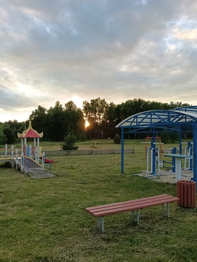 The image depicts an outdoor playground and exercise area at dusk in Smolino, Russia. The scene is set on a grassy field, with a dense line of trees forming the background. The sun is setting behind the trees, casting a warm glow and dramatic rays of light through the overcast sky.

In the foreground, a red wooden bench with metal legs sits on the grass. To the right of the bench is a cylindrical brown trash can. Further into the scene, an outdoor exercise station is visible, featuring various metal equipment with blue and white frames, situated under a blue canopy.

To the left, a colorful playground structure with a red and yellow roof and blue railings is present. A ramp leads up to the structure, suggesting accessibility. A wooden picket fence runs across the middle ground, separating the playground and exercise area from a more distant, possibly athletic field marked with white posts. A small pine tree stands near the fence.

The sky is filled with clouds, indicating a somewhat cloudy evening, with the sunset creating a soft, diffused light. No people are visible in the image. The overall atmosphere is peaceful and serene, suggesting the end of a day. There is no visible text in the image.
