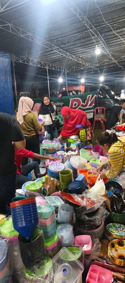 This image depicts an outdoor market stall in Banding Agung, Indonesia. A variety of plastic household items, primarily bowls and containers in various colors, are displayed for sale. Several people, some wearing headscarves, are present at the stall, appearing to be customers and vendors. The stall is covered by a large, tent-like structure supported by metal beams. In the background, a vehicle is visible, along with more market stalls and structures. The lighting suggests it is either evening or nighttime. The overall scene captures a bustling marketplace atmosphere.