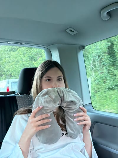 A female passenger sits in a car, holding a grey travel neck pillow, possibly preparing for a trip. The background shows greenery through the car window. This image was taken in Mali Zvornik Municipality, Serbia.