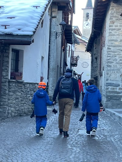 A group of people dressed in winter ski attire walks along a narrow, cobbled street in the mountain town of Courmayeur, Italy. The scene depicts a typical European alpine village, with buildings constructed from stone and plaster, some featuring traditional wooden balconies. Snow-covered rooftops are visible, confirming a cold, wintery environment.

In the foreground, a child wearing a bright blue ski suit and an orange helmet holds a ski pole. Behind them, an adult, dressed in a dark jacket and brown pants, carries a black backpack and also holds a ski pole. Further back, another child in a blue ski suit and ski boots, and a person in a red jacket, are part of the group. They appear to be either en route to or returning from skiing or snowboarding activities.

The street itself is paved with cobblestones, which appear slightly wet, reflecting the light. The time of day is bright, suggesting late morning or early afternoon. In the background, a clock tower with Roman numerals can be seen, with the clock face indicating approximately 10:50 or 10:55. The architectural style, winter conditions, and the subjects' clothing all reinforce the setting of a ski resort town.
