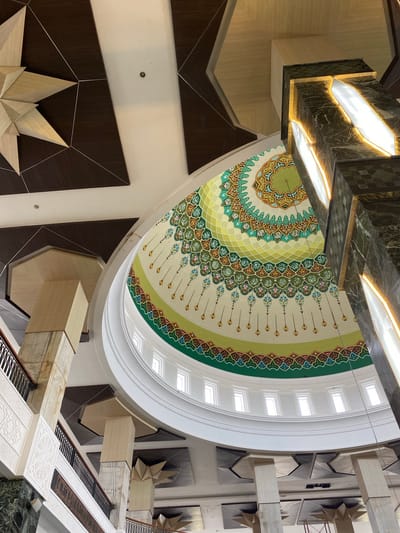 The image shows the interior of a building with a dome featuring intricate geometric and floral patterns in shades of green, yellow, and cream, with Arabic calligraphy integrated into the design. The dome is supported by white columns and a circular white structure with small rectangular windows around its perimeter. To the right, a dark, marble-clad column is illuminated by long, narrow light fixtures. The ceiling above the dome is composed of dark brown wooden panels with silver geometric lines, and a large, wooden starburst-like decorative element is visible. In the lower left and center of the image, there are marble pillars and balustrades of a staircase, with ornamental white carvings on the wall. The scene is well-lit, suggesting it is daytime or the interior is brightly illuminated. The architectural style appears to be modern Islamic, likely a mosque or a similar public building. The specific location of Aluekambuk, Indonesia, is mentioned in the prompt but not directly evident from the image itself, which is a general interior architectural shot. There are no people or animals visible in the image.