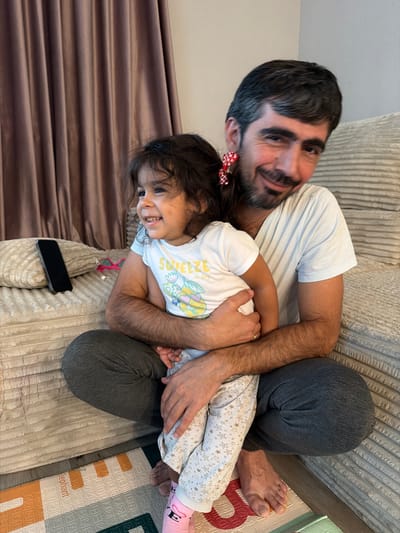 An adult male and a young child are on a sofa. The male is wearing a white t-shirt and gray sweatpants. The child is wearing a white t-shirt with a yellow graphic and patterned pants. Both are smiling and looking in the same direction. The male is holding the child. A phone, a pillow, and a curtain are visible in the background. This image was taken indoors, likely in a residential setting. No specific location within Phoenix, United States of America can be determined from the provided image.