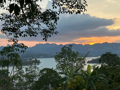 The image is a landscape view from a vantage point in Ban Chiao Lan, Thailand, overlooking Cheow Lan Lake at sunset. The sky is filled with clouds, with the sun setting behind a distant mountain range, casting an orange glow. The lake is visible between dense green foliage, including trees and banana plants. Some of the trees are silhouetted, framing the scene. In the middle distance, there are small islands dotted around the lake. A traffic sign is visible among the foliage. The scene has a tranquil, natural setting.