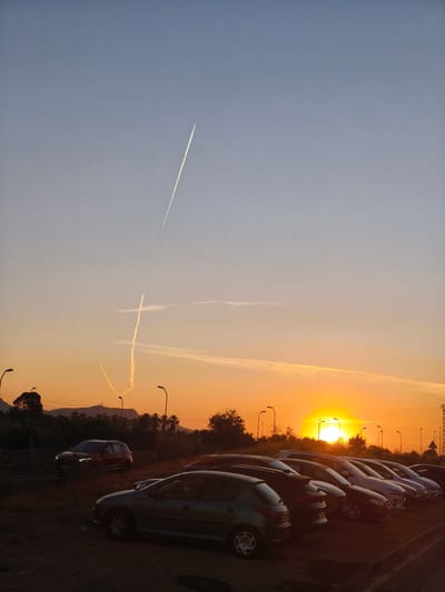 The image captures a parking lot at sunset in Nàquera / Náquera, Spain. Multiple cars are parked in rows, silhouetted against the setting sun. The sky transitions from blue to orange hues. Contrails from aircraft crisscross the sky. Distant trees and streetlights punctuate the horizon.