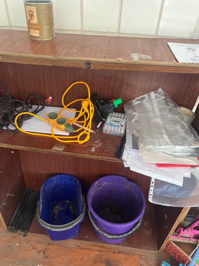 The image shows a shelf in what appears to be a utility or storage area. On the top shelf, there is a can of Turkish coffee, a power strip with a yellow cord, and a collection of markers in a plastic case. There are also some plastic document protectors and papers stacked on the right side of the shelf. The bottom shelf holds two buckets, one blue and one purple, and a stack of black items that could be rods or poles. The environment appears somewhat cluttered and dusty, with some debris on the wooden floor. The wall behind the shelves is tiled with white tiles. The image does not provide any explicit indicators of the city of Mlyniv, Ukraine, or any people.