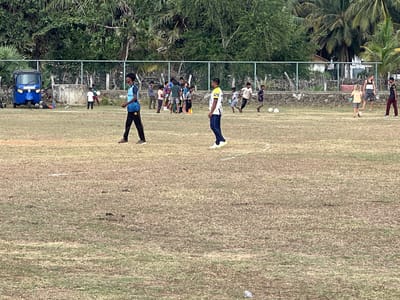 The image depicts a vibrant daytime scene on a dry, grassy field in Ulla, Sri Lanka. Approximately 15-20 individuals, mainly males ranging from young children to young adults, are scattered across the field, participating in what appears to be a casual football practice or game.

In the mid-ground, a young man wearing a light blue and dark blue sports jersey with the number "05" and "SPORT CLUB" visible on the back walks towards the left, while another young man in a yellow and white shirt stands facing right. Several children and young adults are seen interacting with multiple footballs, some running and others standing, suggesting various stages of play or training drills. Orange training cones are also visible on the field.

The background is characterized by a sturdy green metal fence running horizontally, behind which lies a low concrete wall. Beyond the fence, dense tropical foliage, including lush green trees and prominent palm trees, fills the upper background, indicating a warm, natural environment. On the far left, a blue tuk-tuk, a common local vehicle, is parked near the fence, accompanied by bicycles and what appear to be sports bags or additional balls. A building is partially visible in the far-right background, nestled among the trees. The scene conveys a relaxed and active community gathering centered around sport.