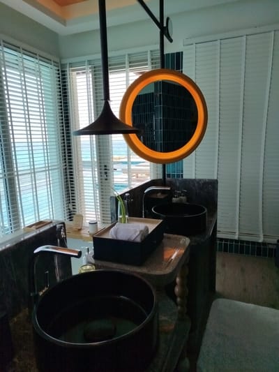 This is a photograph of a bathroom in the city of Kendhoo, Maldives. The bathroom features two dark, round sinks on a marble countertop. Above the sinks, a large, circular mirror with an illuminated orange frame hangs. Behind the mirror, white horizontal blinds cover a large window that offers a view of the ocean and a swimming pool area. There are two faucets, soap dispensers, and a small wooden tray with white towels on the countertop. A dark, cone-shaped pendant light hangs from the ceiling, positioned above the sinks. To the right of the sinks, a light-colored stool is partially visible. The overall impression is one of a luxurious and modern bathroom with a strong connection to the surrounding tropical environment.
