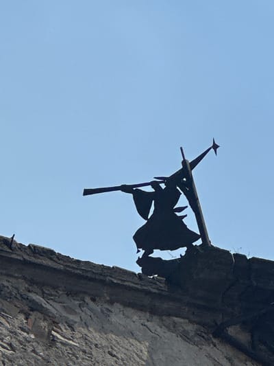 The image shows a close-up of an ornate, dark-colored metal structure attached to the corner of a weathered stone or concrete building against a clear, light blue sky. The metal structure features a prominent star-shaped element atop a rod, which is attached to a horizontal beam. Below the beam, there is a tattered, flag-like metal piece with irregular edges. The building's corner appears to be aged and crumbling, with visible surface texture. The location is presumed to be in Tlalnepantla, Mexico.