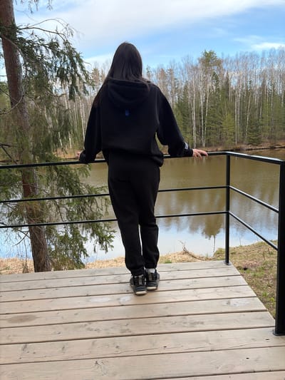 An outdoor, eye-level shot captures a person standing on a wooden deck, leaning against a black metal railing, with their back to the viewer. The person is dressed in a black hooded sweatshirt and black pants, with dark hair cascading down their back. They wear black and white sneakers.

The scene unfolds in a tranquil natural setting. Beyond the railing, a calm body of water stretches out, reflecting the sky and distant trees. The opposite bank of the water is lined with a dense forest featuring a mix of tall, slender birch trees (many with bare branches, suggesting early spring or late autumn) and dark green coniferous trees. The sky above is light blue with sparse, wispy white clouds. The wooden deck beneath the person's feet is made of light-colored planks, showing some signs of wear and knots.

The person appears to be observing the scenic landscape. A small white logo or text is visible on the left sleeve of their hoodie, and a small blue logo, possibly a stylized "H" or a house icon, is visible on the back of the hoodie. The lighting suggests it is daytime, likely during a clear, bright period. The overall atmosphere is one of calm contemplation in nature, consistent with a location like Mishkino, Russia.