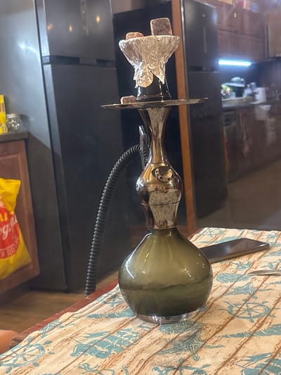 The image shows a hookah on a table in what appears to be a domestic setting in Kamenar, Bulgaria. The hookah is made of glass and metal, with a black hose. There are charcoal pieces on top of the hookah. A smartphone and a fork are also visible on the table. The background includes a refrigerator and kitchen cabinets. The overall lighting suggests an indoor environment.