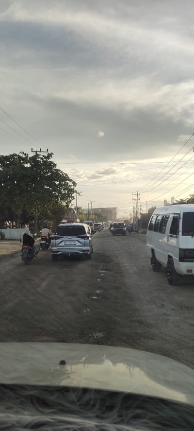 The image depicts a traffic scene on a dusty, unpaved road in Barokulamgajah, Indonesia. The sky is overcast with grey clouds, and the lighting suggests it is either early morning or late afternoon. In the foreground, the hood of a vehicle is visible, reflecting the surroundings.

The road is lined with electrical poles and wires. To the left, a large tree with dense green foliage is prominent. On the road, there is a queue of vehicles. A silver SUV is in the middle ground, facing away from the camera, with a license plate that reads "BL 1762 ZN". To its left, a person wearing a hijab is riding a scooter, also facing away. Further ahead, several other vehicles, including cars and what appears to be a white van or minibus, are present in the traffic jam. The road surface is uneven and shows signs of water pooling in some areas, indicating it is muddy or has potholes. Buildings and structures are visible in the distance on the right side of the road. The overall atmosphere suggests a typical day with traffic congestion in a semi-rural or developing urban area.