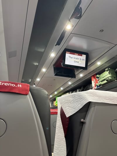 The image captures the interior of a modern train carriage, specifically an Italian high-speed Italo train, indicated by the "italo" logo on a display and a "treno.it" fabric tag. The scene is taken from a tilted, low-angle perspective looking upwards and along the carriage.
The setting is well-lit by numerous small LED lights embedded in the ceiling and along the overhead luggage racks. The interior features a clean design with grey and white surfaces. A digital screen on the left displays "italo", the number "8158", the current time "20:07", and the message "The train is on time", signifying an evening journey that is running punctually.
On an overhead luggage rack, a small, light grey suitcase and a red-and-white striped bag or item are stored. To the right, a large amount of crumpled white tissue paper is draped over what appears to be the top of a seat or partition. A red fabric tag, clearly visible on a seat, reads "treno.it" and partially "italotren". Seats are upholstered in a dark grey material with subtle red or burgundy accents. The overall impression is one of a calm and orderly train journey through Italy, with the time of day being evening.