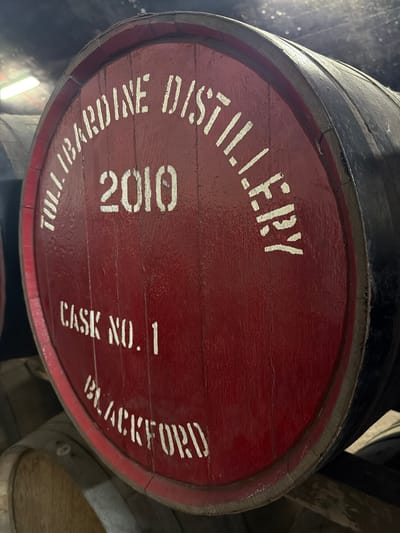 The image shows the end of a whiskey barrel with red paint and white lettering. The inscription on the barrel reads "TULLIBARDINE DISTILLERY," "2010," "CASK NO. 1," and "BLACKFORD." The setting is likely a warehouse or storage facility where barrels are stacked, as other barrels are partially visible in the background. Blackford is a town in Scotland, United Kingdom.