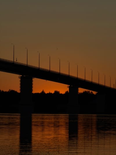 A silhouetted bridge spans across the frame, its structure dark against an orange sunset sky. The bridge has a railing and is lined with evenly spaced streetlights. Below the bridge, a dark band of trees forms a silhouette along the horizon. The water in the foreground reflects the warm colors of the sky and the dark structure of the bridge. There are no people visible in the image. The scene appears to be a landscape at dusk, likely near Tarasovo, Russia, given the provided context.