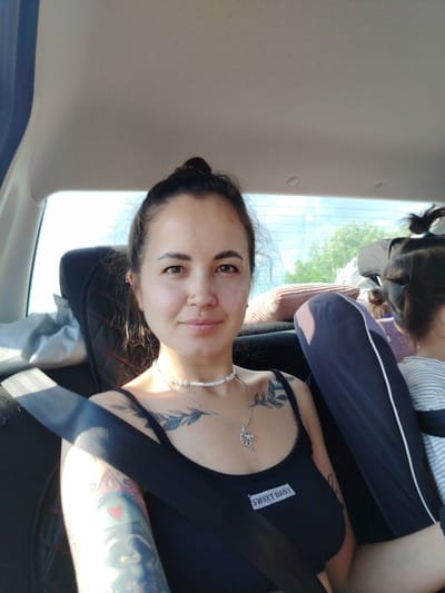 A woman with dark hair pulled into a bun and multiple tattoos is taking a selfie in the back seat of a car. She is wearing a black tank top and a pearl necklace. A young child with dark hair in pigtails is seated next to her. The car is likely in motion as indicated by the seatbelt. The background shows trees and a blue sky, suggesting an outdoor setting, possibly on the outskirts of the city of Bol'shoy Gondyr', Russia.