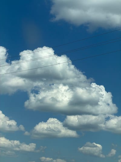 This is a vertical, low-angle, eye-level shot of a bright blue sky with fluffy white clouds. Four dark, thin electrical wires diagonally traverse the image from the bottom left to the top right. The wires are strung close together and appear taut. The clouds are voluminous and dynamic, with some areas of shadow, suggesting significant depth. The sun is not directly visible, but its bright light illuminates the clouds, creating a contrast between brightly lit portions and shadowed undersides. The overall impression is one of a clear, breezy day. The image was taken outdoors, likely in a rural or suburban setting given the presence of overhead electrical wires. The sky and cloud formations suggest it is daytime, likely midday or afternoon due to the brightness and the shadows on the clouds. There is no visible text in the image. The lack of any discernible man-made structures other than the wires, combined with the expansive sky, creates a sense of openness and nature.