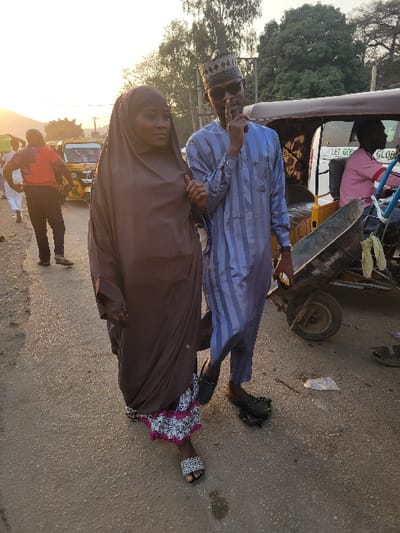 A man in a blue striped shirt and traditional cap stands next to a woman in a brown hijab on a street. The woman wears sandals. A yellow vehicle is behind them. Another man in a red shirt is seen in the background. A wheel and miscellaneous items are visible near the first man. The scene appears to be outdoors with a road surface in view.