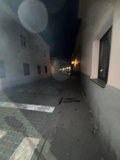 The image displays a narrow, unpopulated street at night in Sibiu, Romania. The scene is poorly lit, with light originating from distant streetlights. The ground consists of cobblestones in the foreground on the left, exhibiting faded white pedestrian crossing stripes. This transitions to a darker, smoother paved surface further into the middle ground. A prominent dark reddish-brown, irregular stain, consistent with dried blood, is present on the paved surface, slightly right of the center, accompanied by several smaller dark specks.
Buildings flank both sides of the street. The building on the right features a light-colored upper facade and a darker, possibly grey or blue, lower half. A dark, rectangular window with a prominent sill is visible on this building. The building on the left is a light-colored, multi-story structure with several dark, rectangular windows.
The street extends into the background, where a series of orange streetlights are visible. A dark-colored vehicle is dimly discernible further down the street, facing away from the viewer. No individuals are present in the image.