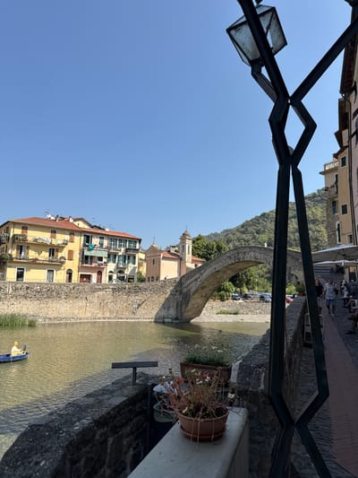 The image depicts a scenic view of the Nervia River in Dolceacqua, Italy, on a clear, sunny day. In the foreground, a dark metal structure with a square lantern is visible on the right, partially obscuring the view of a stone wall and a sidewalk. Several potted plants with red and white flowers sit on the wall. The river flows calmly, with its clear, shallow water reflecting the sunlight. A small blue boat with a person wearing a yellow hat and holding a black object is visible on the left side of the river.
The iconic Ponte dei Cavalieri (Knights' Bridge), a medieval stone arch bridge, spans the river. On the far bank of the river, a row of colorful buildings with red tile roofs lines the embankment. To the left of the bridge, a church with a bell tower is visible, and beyond that, a lush, green hillside rises. On the right side of the bridge, a paved walkway with outdoor seating for a cafe or restaurant is present, with a few people walking and sitting. The overall atmosphere is tranquil and picturesque, showcasing the charm of this Italian town.