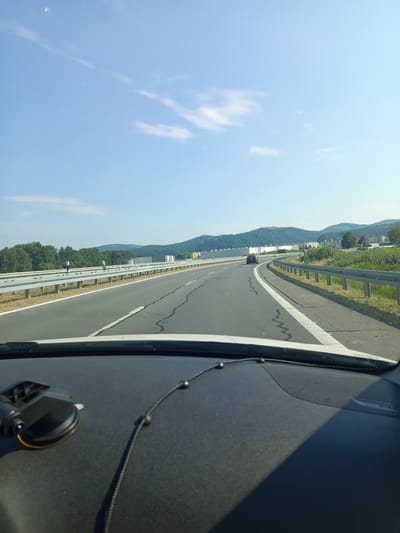 The image depicts a view from the driver's seat of a vehicle traveling on a highway. The road ahead curves to the right, with a silver guardrail separating the lanes. The highway is flanked by green trees and rolling hills in the distance. Several buildings, possibly industrial or residential, are visible on the right side of the road. The sky is a clear blue with a few wispy white clouds. The foreground shows the dashboard of the car, with a black suction cup mount on the left and a black cord with small spherical objects running across it. The license plate of a dark-colored car is visible in the distance on the highway. The highway is identified as E77 in the Zvolen region of Slovakia.