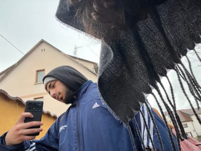 The image captures a low-angle perspective of a young man holding a smartphone in Meistratzheim, France, under an overcast sky. The man, positioned slightly left of center, has a beard and is wearing a grey beanie beneath a dark blue hooded jacket featuring white Adidas stripes on the sleeves and an Adidas logo on the chest. He holds a black smartphone with a distinctive multi-lens camera module in his right hand, angled upward, with his gaze directed toward the screen. The upper right portion of the frame is partially obscured by a dark, textured fabric with fringes. In the background, residential buildings are visible, including a light beige house with a prominent gable roof, two small rectangular windows, and a television antenna. Patches of ice or snow are visible along the eaves of the houses, and bare tree branches are discernible against the cloudy sky. A wire or cable traverses the upper background horizontally.