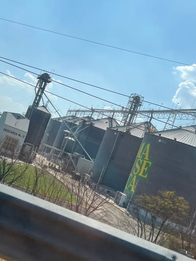 A view of a grain elevator complex, featuring tall, dark silos with steel support structures against a clear blue sky with scattered clouds. Green and yellow lettering is visible on one silo. Power lines cross the frame. Adjacent to the silos is a small building marked "DESCARGA No. 2." Grass and trees are on one side of the silos. A gray object appears partially in the right foreground, possibly part of a window or structure from where the photo was taken.