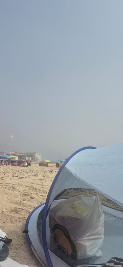 The image shows a beach scene in Póvoa de Varzim, Portugal, with a blue and white beach tent in the foreground. Inside the tent, a white plastic bag with green and orange labeling is visible, containing what appears to be food items or groceries. The bag is labeled "REUTILIZAVEL" and "270%". The tent is pitched on a sandy beach, with other beachgoers and umbrellas in the background. A yellow flag is planted on a pole to the left of the frame, and a building complex is situated further back, partially obscured by a light fog. The sky is overcast with a grayish-blue hue. The sand is textured with scattered debris. To the left of the tent, a black sandal and a white shoe are visible on the sand.