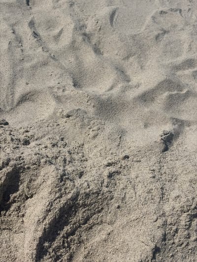 This is a close-up, high-angle, outdoor, daytime photograph of granular beach sand. The sand is a light brown and grey color, with visible texture and ripples created by wind or water. There are no people, objects, or discernible human activity in the frame. The image focuses solely on the natural patterns and textures of the sand. This scene is consistent with a beach environment, such as the coastline of Povarovka, Russia, a location known for its sandy shores along the Gulf of Finland.
