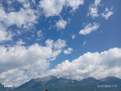 The image depicts a panoramic view of a mountain range under a bright blue sky filled with fluffy white clouds. The lower portion of the image is dominated by the verdant, tree-covered slopes of the mountains, with jagged, rocky peaks rising above. The sky is a vibrant blue, with scattered clouds ranging from thin, wispy formations to larger, cumulus-like clouds. The overall impression is one of a clear, bright day in a mountainous region. A timestamp and device model are present in the bottom corners of the image: "REDMI 13C" on the left and "16.07.2026 11:13" on the right. No people or specific city landmarks are visible, but the description indicates the location as Khurai-Khobok, Russia.