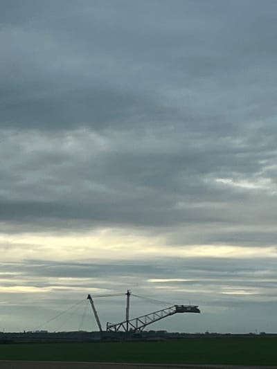 The image depicts a vast, flat landscape under an overcast sky in Jüchen, Germany. A large, multi-component industrial machine, consistent with a bucket-wheel excavator or similar opencast mining equipment, dominates the midground in silhouette. The machine features a complex truss structure, including an prominent, angled boom extending to the right from a central support framework. A tall, slender mast is visible on the left side of the machine, with cables extending outwards. The machine is stationary. The sky occupies approximately the upper two-thirds of the frame, filled with dense, grey clouds, with a horizontal band of lighter, yellowish-grey clouds visible near the horizon line. The lower third of the image consists of dark green, flat terrain, indicative of cultivated fields or reclaimed land. No human figures or active operations are visible.