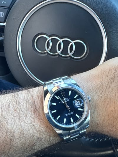 The image shows a man's hairy wrist wearing a Rolex Oyster Perpetual watch, with a dark blue dial and a date indicator at the 3 o'clock position. The watch has a stainless steel bracelet and case. In the background, partially obscured by the arm and watch, is the steering wheel of an Audi, identified by the four interlocking rings logo. The lighting suggests the photo was taken indoors, likely inside the car, possibly during daytime. There is no indication of the image being taken in Krichim, Bulgaria.