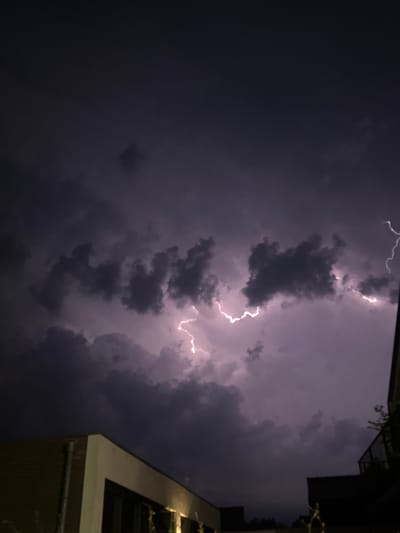 An atmospheric lightning strike illuminates dark, turbulent storm clouds over the city of Putten, Netherlands. In the foreground, the dark silhouettes of buildings are visible, with the corner of one brick building catching some of the ambient light. The lightning, a bright white jagged line, cuts through the purple-tinged clouds, highlighting their texture and density. The scene captures a moment of intense weather activity.