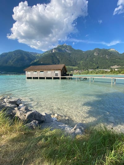 This media file captures a picturesque waterside scene on a bright, sunny day. The primary subjects include a wooden boathouse situated on the edge of a clear, turquoise lake or river. The boathouse has a distinctive sloped roof and several windows visible on its side, supported by wooden stilts that extend into the water. Beyond the water, a lush, green mountainside dominates the background, dotted with trees and some scattered buildings suggesting a small village or resort. The sky is a vibrant blue, with a few fluffy white clouds adding depth to the scene.

The water itself is shallow near the shore, with rocks and patches of submerged vegetation visible through the transparent surface. Sunlight reflects off the water, creating dappled patterns. To the right of the frame, a grassy bank with tall green grass meets the rocky shoreline. There are no people visibly active in the foreground or near the boathouse, suggesting a quiet moment. A long wooden pier extends from the shore into the water, with several supports visible, and two figures can be faintly seen near the end of the pier.

The overall impression is one of tranquility and natural beauty, likely captured during the daytime given the bright sunlight and clear skies. The setting appears to be a serene natural environment, possibly a lakeside or riverside area in the Austrian Alps, given the mention of Lärchenwiese, Austria. The weather is ideal for outdoor activities, with the sun casting shadows and illuminating the landscape. No visible text is present in the image.