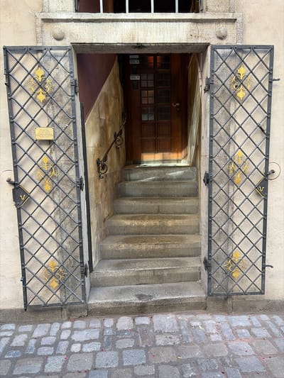 The image shows an exterior view of an entrance to a building in Würzburg, Germany. The entrance is recessed and approached by a set of stone stairs. The stairs lead up to a wooden door with glass panes. On either side of the entrance are large, ornate metal grilles with a diamond pattern, each adorned with a golden fleur-de-lis design. The grilles are open, revealing the entryway. A sign with text in German is attached to the left grille. The ground in front of the entrance is paved with cobblestones. The walls of the building are a light, textured material, and the architectural details suggest an older, possibly historic, structure. There are no people visible in the image.