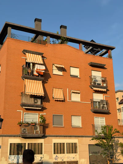 This media file captures a view of an apartment building in Benimàmet-beniferri, Spain, likely taken from a higher vantage point, possibly another building or drone. The building's facade is constructed with red bricks, and it features multiple windows, some with awnings. Several windows have metal grilles or security bars, and some are equipped with air conditioning units. There are also balconies with some greenery, suggesting residential occupancy.
The scene depicts the exterior of the building under a clear blue sky, indicating a sunny day. The sunlight casts shadows on the brickwork, suggesting it is either morning or late afternoon. The composition is slightly tilted, giving a dynamic perspective of the architecture.
On the far right of the image, a portion of another building is visible, which appears to have a lighter-colored facade and larger windows, some adorned with graffiti. A person's head is visible in the bottom right corner, looking towards the building.
One of the windows on the brick building has laundry hanging on a rack, and another has a blanket draped over it, implying everyday life activities within the apartments. The overall impression is a slice of urban residential life in Spain, characterized by functional architecture with decorative elements like awnings and security features. No discernible text is visible in the media file, aside from what appears to be graffiti on the adjacent building.