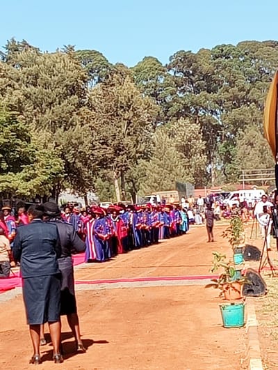 This media file depicts an outdoor formal event, likely an academic procession or ceremony, taking place under a clear blue sky in Kimumu ward, Kenya.
The main subjects are a long line of individuals, presumably academics or graduates, dressed in distinctive academic regalia. They wear vibrant gowns, predominantly blue and red, with visible hoods and mortarboards or caps, suggesting a graduation or convocation event. They are seen moving in an orderly fashion along a wide, unpaved pathway of reddish-brown earth.
In the foreground, two women in dark, knee-length dresses or uniforms stand with their backs to the camera, appearing to observe or manage the event. The pathway features a narrow red carpet-like runner down its center, adding to the ceremonial atmosphere. Along the right side of the path, potted green plants and black speakers or sound equipment are arranged, indicating an organized setup for an audience or address. Further in the background, several other people are scattered, some standing by the pathway and others near parked vehicles, including vans and trucks.
The setting is characterized by a dense backdrop of numerous tall, lush green trees that fill the mid-ground and background, suggesting a natural or park-like institutional campus environment. The bright, clear sunlight of what appears to be mid-morning or early afternoon casts visible shadows on the ground. The overall scene conveys a sense of celebration and formality. No visible text is legible within the image.