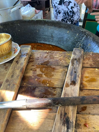 The image shows a close-up view of a large pot filled with a reddish-brown stew, likely being prepared outdoors in Meunasahmanyang, Indonesia. The pot is positioned over a wooden surface, which is covered in stains and debris, suggesting a food preparation area. Several wooden planks are visible on the surface, and a metal utensil with a wooden handle rests on top of them. In the background, a person wearing a patterned shirt is visible, along with other food preparation items and containers. The lighting suggests it is daytime, and the scene appears to be a busy outdoor market or a street food stall.