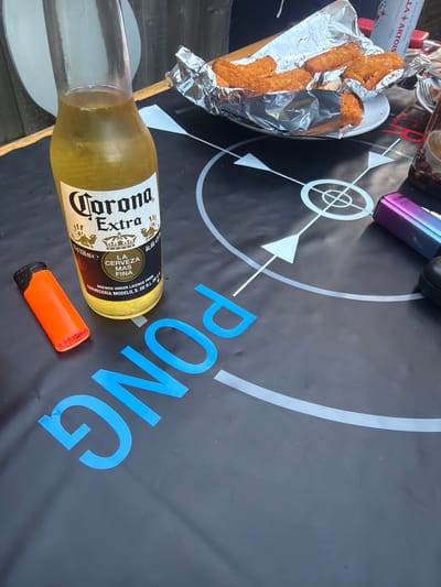 The scene depicts a partially cropped table with a black tablecloth printed with a white beer pong design. A half-empty bottle of Corona Extra beer lies on the tablecloth, near an orange highlighter and the blue text "PONG" printed on the table covering. A plate containing fried food wrapped in aluminum foil is visible in the upper left. Part of a Stella Artois can and an unidentifiable device are also on the table. The location is Swindon, United Kingdom. There are no people visible in the image.
