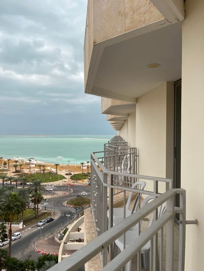 The image captures an elevated perspective from a building, likely a hotel, looking out over a coastal scene. In the immediate foreground on the left, a light-colored concrete balcony structure with a recessed ceiling light is visible. This architectural style, featuring repetitive angular elements, extends into the distance along the side of the building.
To the right, a metal railing, likely part of another balcony, frames the view. Beyond it, a road with a roundabout can be seen, populated by a few cars, some parked and others in motion. The road is flanked by green landscapes featuring numerous palm trees. Further along, a sandy beach is visible, adorned with multiple white canopy structures or sun loungers.
The background is dominated by a serene body of water, distinguished by its striking turquoise hue, which stretches to the horizon under an overcast sky filled with light grey clouds. The unique color of the water is a strong indicator of the Dead Sea in Ein Bokek, Israel. The time of day appears to be daytime, though the cloudy conditions diffuse the light. No people are prominently visible, and no discernible text is present in the image. The overall atmosphere is calm and expansive.