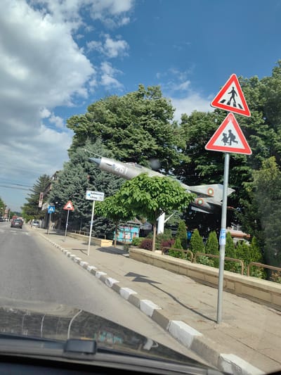 The image is taken from inside a vehicle, looking out onto a street in Yoakim Gruevo, Bulgaria. On the right, a pedestrian walkway is lined with a low stone wall and greenery. Two red triangular warning signs are mounted on a single pole: one indicates a pedestrian crossing, and the other warns of children. A silver jet fighter, marked with the number "261" and roundels, is mounted on a pillar as a monument. Street signs in Cyrillic and English ("Library") are visible. On the left, a street with cars and buildings runs into the distance. The sky above is blue with scattered clouds.
