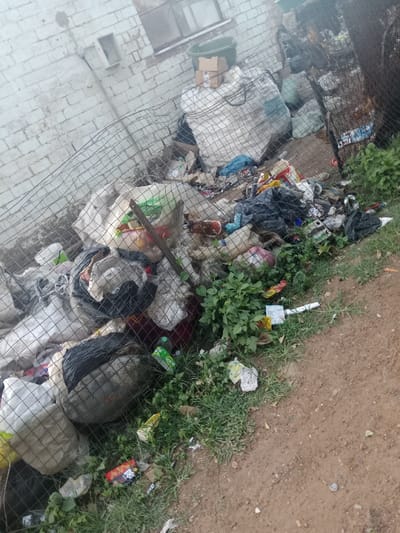 The image captures a large accumulation of garbage and refuse piled behind a wire mesh fence in an outdoor setting in Soweto, South Africa. The scene is dominated by a substantial heap of waste, primarily consisting of plastic bags, various bottles, food wrappers, cardboard, and general household debris. Several large, off-white mesh sacks, possibly filled with recyclable materials or more trash, are prominent, one of which has a cardboard box and a green plastic bucket resting on top.
The waste extends from against a white brick wall of a building, stretching along the fence line, and spilling onto the ground in the foreground. The building features small, rectangular window openings. The fence, a dark wire mesh, acts as a barrier, though some smaller pieces of litter are visible on the outside of it, mingled with weeds and sparse grass. The ground in the foreground is a dirt path with patches of green vegetation.
No human subjects or direct activity are visible, but the scene implies a significant issue of waste management and environmental pollution. The lighting suggests it is daytime, possibly overcast or hazy, as there are no sharp shadows.
Visible text includes "JAVA" on a red and black wrapper near the bottom center, and faint, illegible text on a small green carton that appears to be a juice box or similar container. Small, unreadable labels are also present on the cardboard box resting on the large white sack. The image portrays a neglected area facing substantial sanitation challenges.