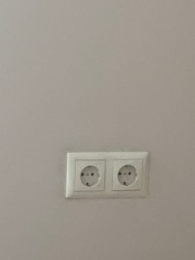 The image shows a close-up view of a dual electrical outlet mounted on a light grey wall. The outlet is white and has two socket holes, each with three circular openings for prongs. The wall appears to be a plain, textured surface. The image is a still photograph and does not contain any information about people, actions, interactions, or the specific location within Ventspils, Latvia. The lighting is even, and there are no shadows that would indicate the position of a light source.