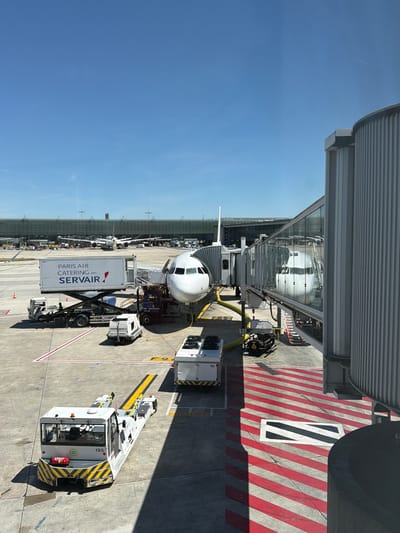 An aerial view of an airport tarmac at Le Mesnil-Amelot, France, on a clear day. A commercial airplane is parked at a gate, connected to a jet bridge. Several ground service vehicles, including a catering truck and a pushback tractor, are positioned around the aircraft. In the background, other aircraft and airport buildings are visible. The tarmac is marked with yellow and white lines, and red and white striped safety zones.
