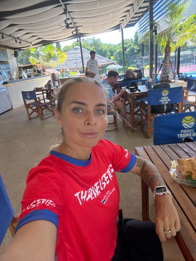 A woman with blonde hair and blue eyes takes a selfie in an outdoor cafe. She is wearing a red t-shirt with blue trim, and a tattoo is visible on her arm. She is sitting at a wooden table, and a pastry is on a plate next to her. In the background, other people are seated at tables, and palm trees are visible. The cafe appears to be part of a Padel Tropical Club, indicated by the blue chairs with the club's logo. The location is identified as Ban Bang Rak, Thailand.
