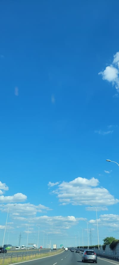 The image depicts a bright, clear day with a vivid blue sky filled with scattered white clouds. The perspective is from a moving vehicle, likely on a highway, as indicated by the road, lane markings, and a line of vehicles stretching into the distance. Streetlights line the side of the road at regular intervals. The right side of the image shows a highway with several cars and trucks traveling in the same direction. A gray car is prominent in the foreground on the right. The scene suggests a daytime journey, possibly during summer or a clear spring day given the bright sunlight and the nature of the clouds. The overall impression is one of travel and open road under a beautiful sky. There is no visible text in the image.